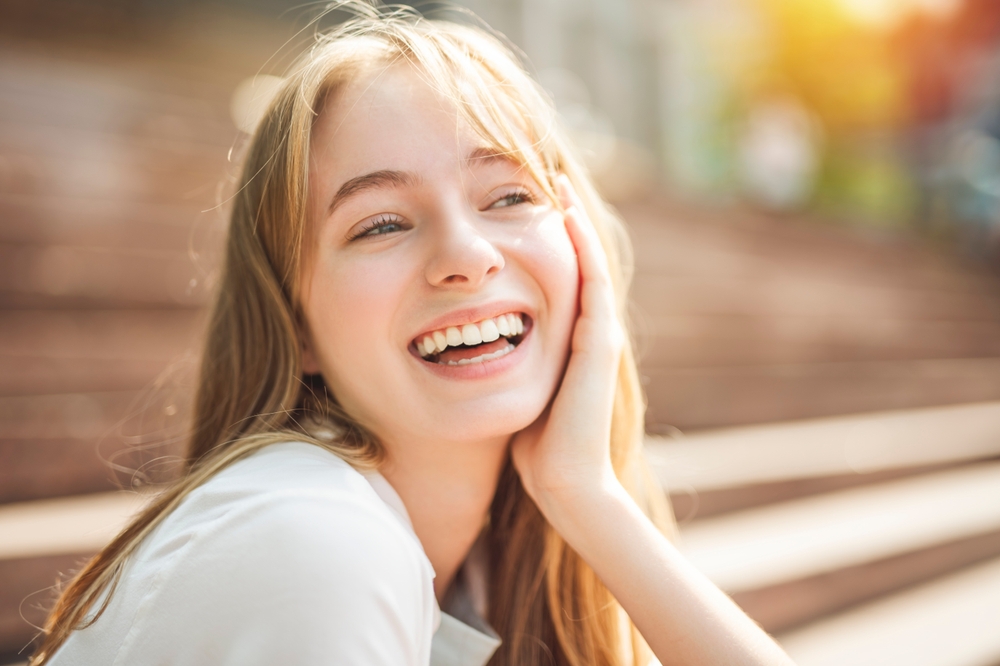 Happy young woman sitting outside with her hand on her cheek, showing a radiant, white smile - Teeth Whitening.