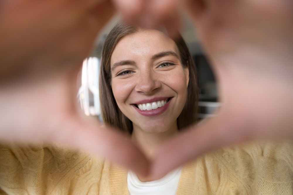 Woman Smiling After Dental Treatment – Minimal Preparation Restorative Techniques A happy woman forms a heart shape with her hands while showing a bright, healthy smile, symbolizing the beauty and confidence restored through minimal preparation restorative techniques.