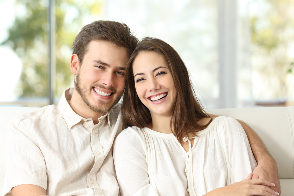 Happy couple smiling while sitting together on a couch - Cosmetic Dentistry Kingsport Young couple sitting close on a couch, both smiling brightly in natural light, wearing light-colored shirts - Cosmetic Dentistry Kingsport