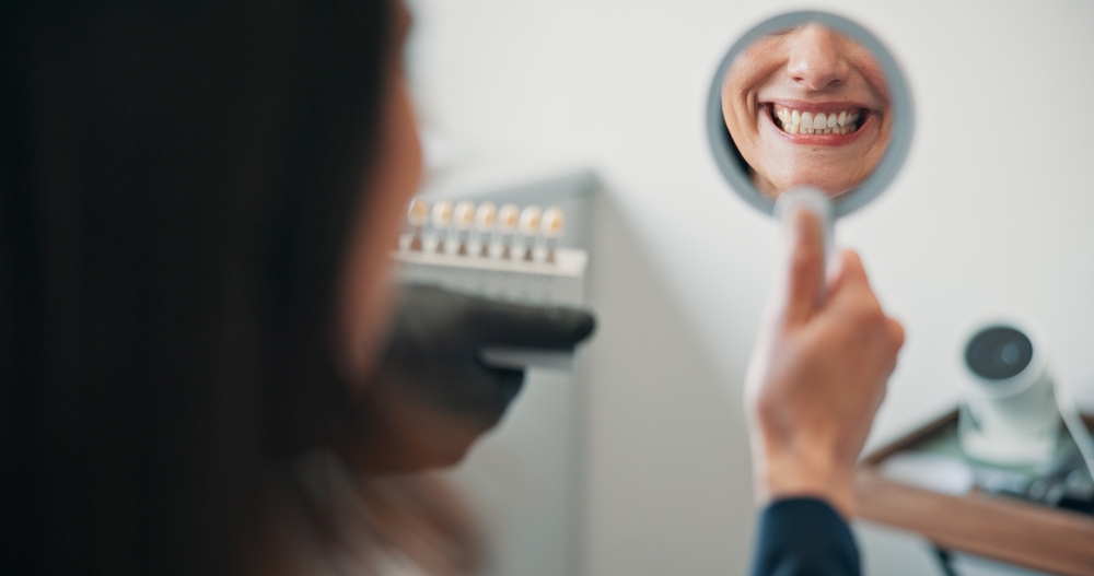 Patient Smiling in Mirror After Dental Restoration - Implant-Supported Crowns and Bridges A patient admires their bright smile in a handheld mirror after receiving dental restoration, showcasing the beauty and precision of implant-supported crowns and bridges.