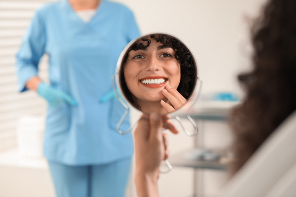 Patient Admiring Smile in Mirror - Porcelain Crowns and Bridges A happy patient looks into a handheld mirror, admiring her bright, restored smile after receiving porcelain crowns and bridges for a natural, flawless look.