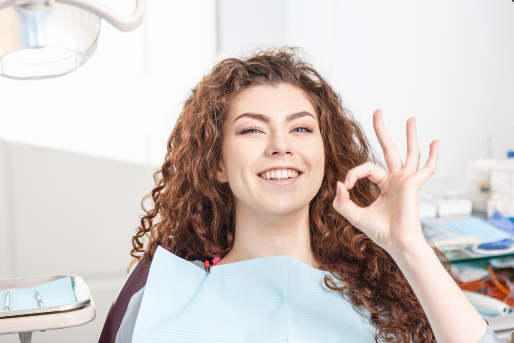 Happy Patient After Dental Cleaning - Comprehensive Dental Hygiene Program A smiling woman gives an OK hand gesture while sitting in a dental chair, showing confidence after receiving a professional cleaning – Comprehensive Dental Hygiene Program.