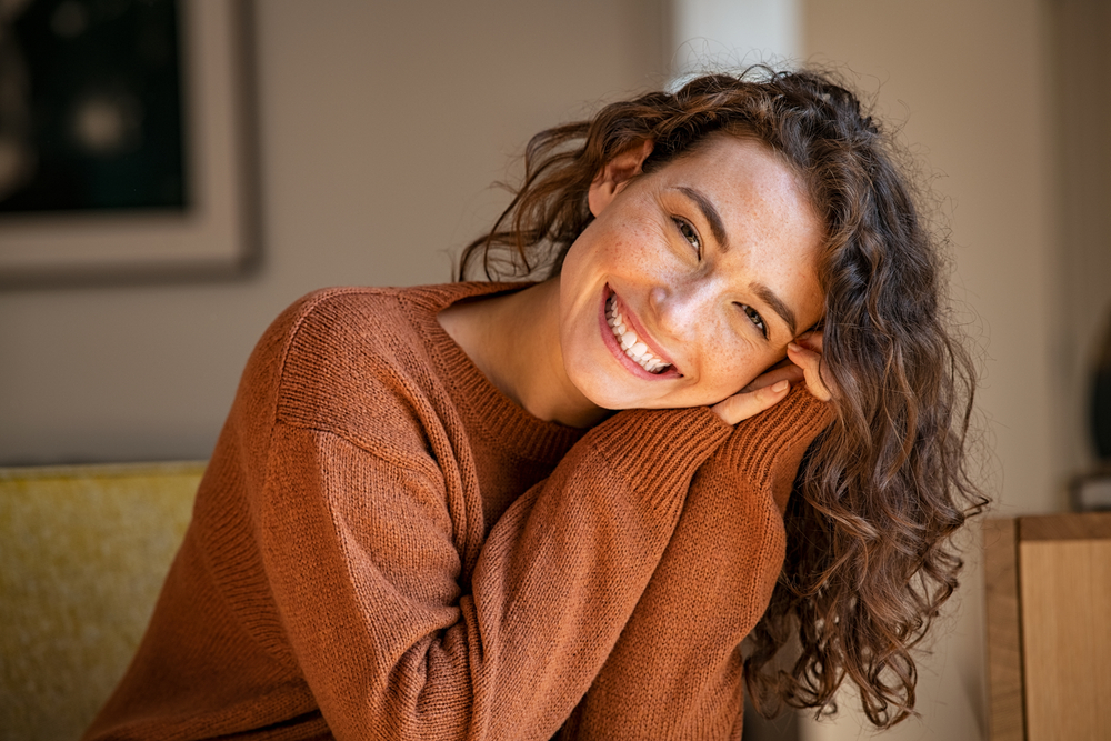 Smiling woman with bright white teeth indoors - Cosmetic Dentistry Kingsport Young woman with curly hair wearing a brown sweater, smiling brightly and showing her white teeth - Cosmetic Dentistry Kingsport